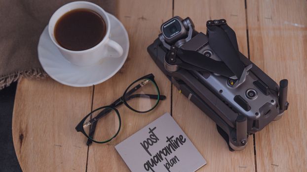 From above of coffee cup and eyeglasses placed on wooden table near folded drone and sticky note with post quarantine plan inscription