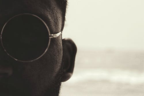 Free Closeup of crop unrecognizable young bearded male in stylish round sunglasses standing against waving ocean on sunny day Stock Photo