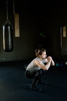 A young man squats in a dimly lit gym, focusing on fitness and strength training with a punching bag nearby.