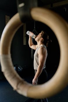 Caucasian man drinking water in a gym setting, framed by gymnastic rings.