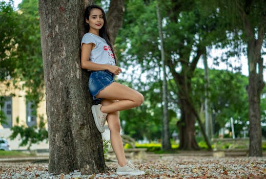 Full body side view of cheerful teen girl in casual clothes leaning on tree trunk in park