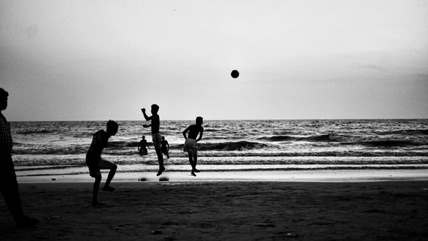 Silhouetted boys joyfully playing soccer on a Mumbai beach in black and white.