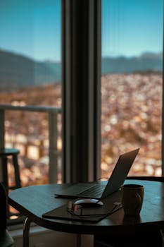 A laptop and mug on a work desk by a window with a scenic city view.