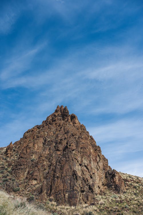 From below scenery view of dry rough mount with uneven surface and pointed peak under bright blue sky with clouds in afternoon