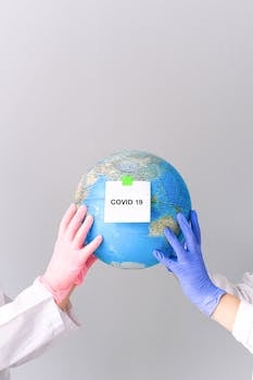 Hands in gloves hold a globe with 'COVID-19' label on a gray backdrop, symbolizing pandemic impact.