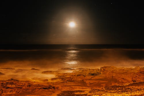 Picturesque view of glowing sun in sky illuminating endless ocean and rough mounts at night at bright sunset