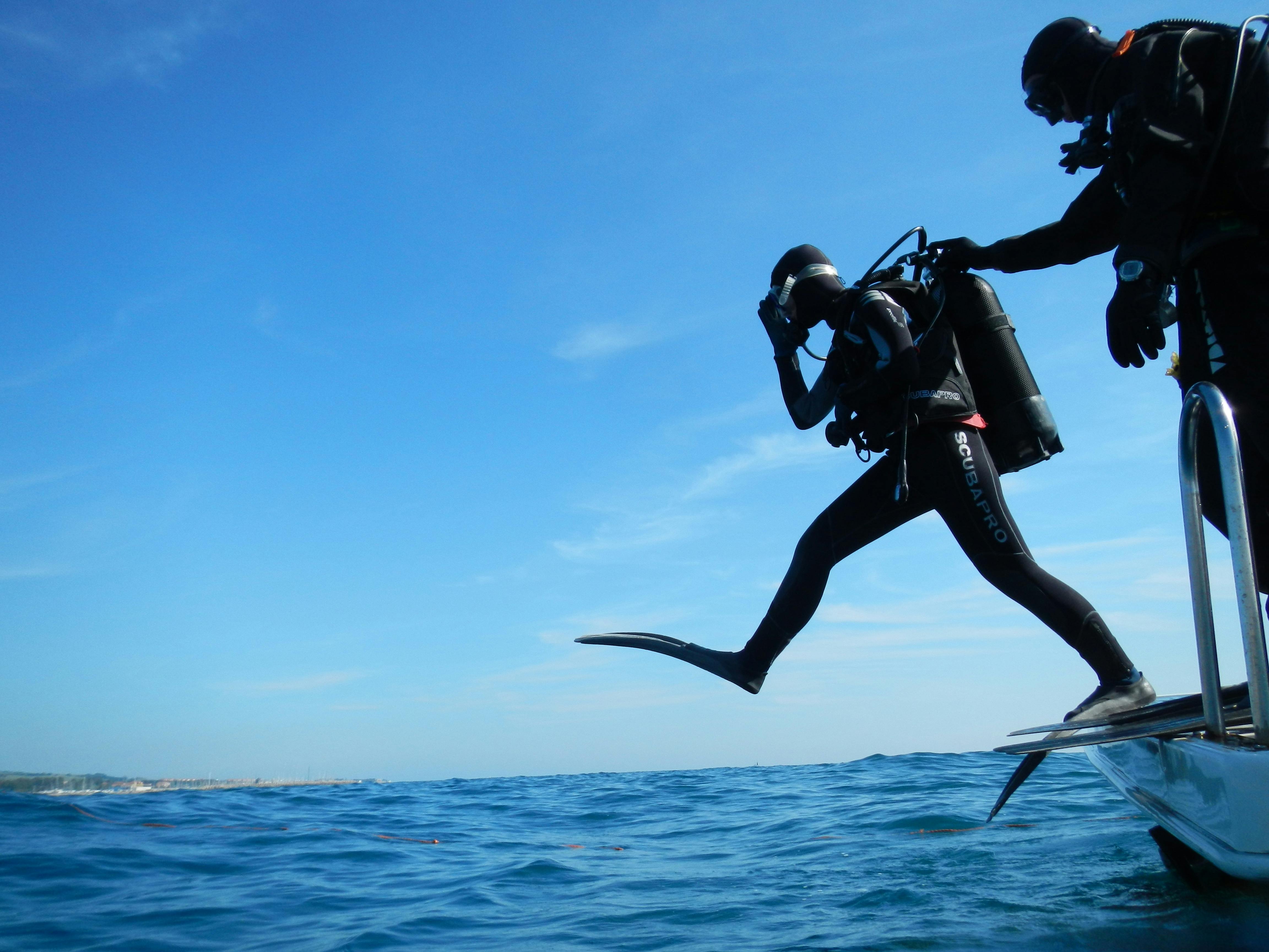 Two divers preparing to step off the edge of a boat. Both are dressed head to toe in scuba gear, one is already stepping out, it appears as if the other may be pushing the other's shoulder.