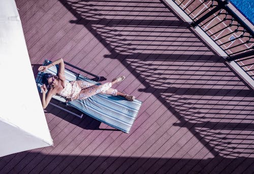 Top view of anonymous female traveler in sunglasses lying with crossed legs on sun bed on wooden floor near fence of hotel balcony in sunlight during trip