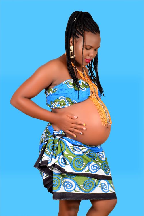 Free Side view of African American expectant female in colorful wear and accessories with makeup touching big tummy while standing near blue wall Stock Photo