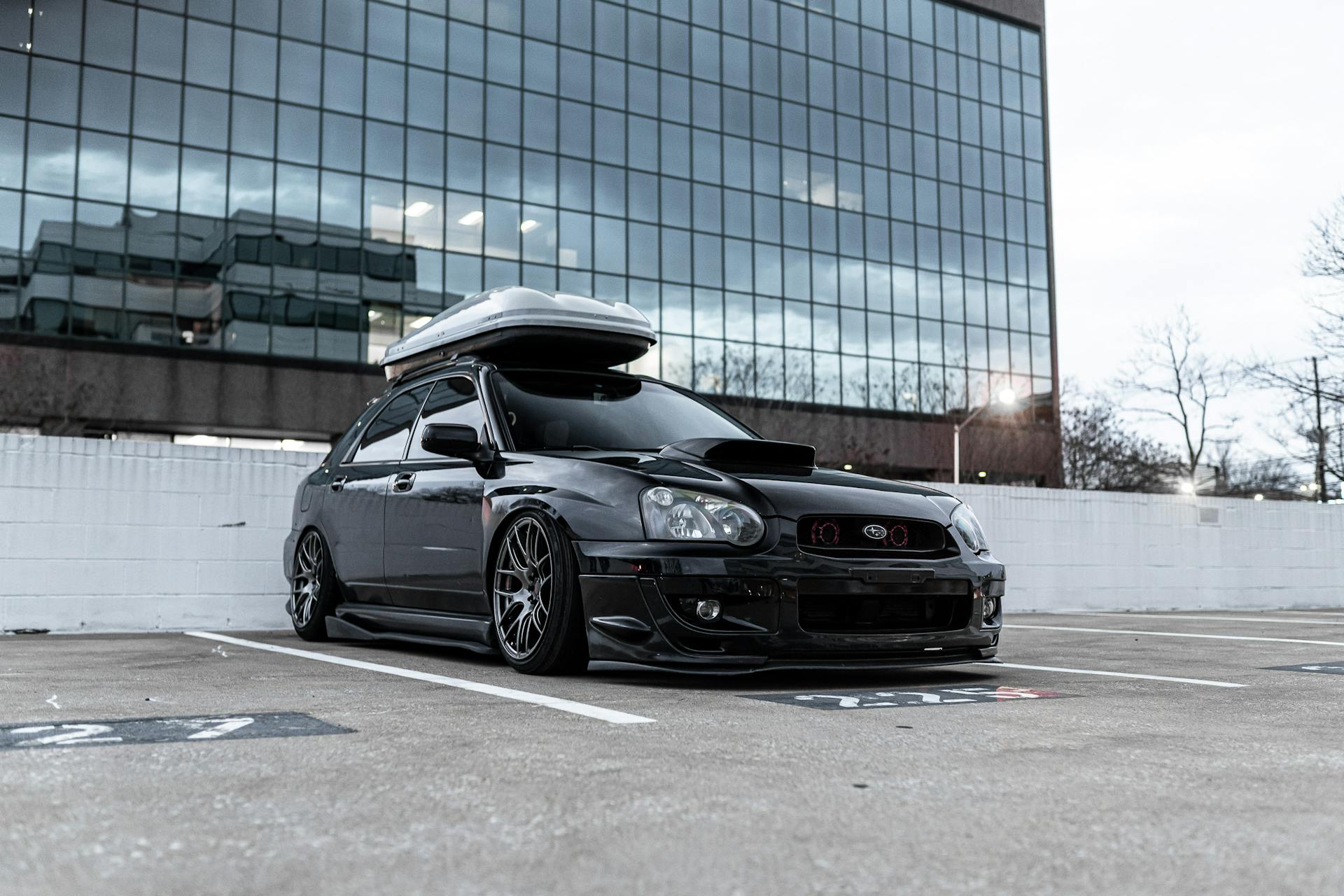 A sleek black car with a luggage rack, parked in an urban parking lot in front of a modern glass building.