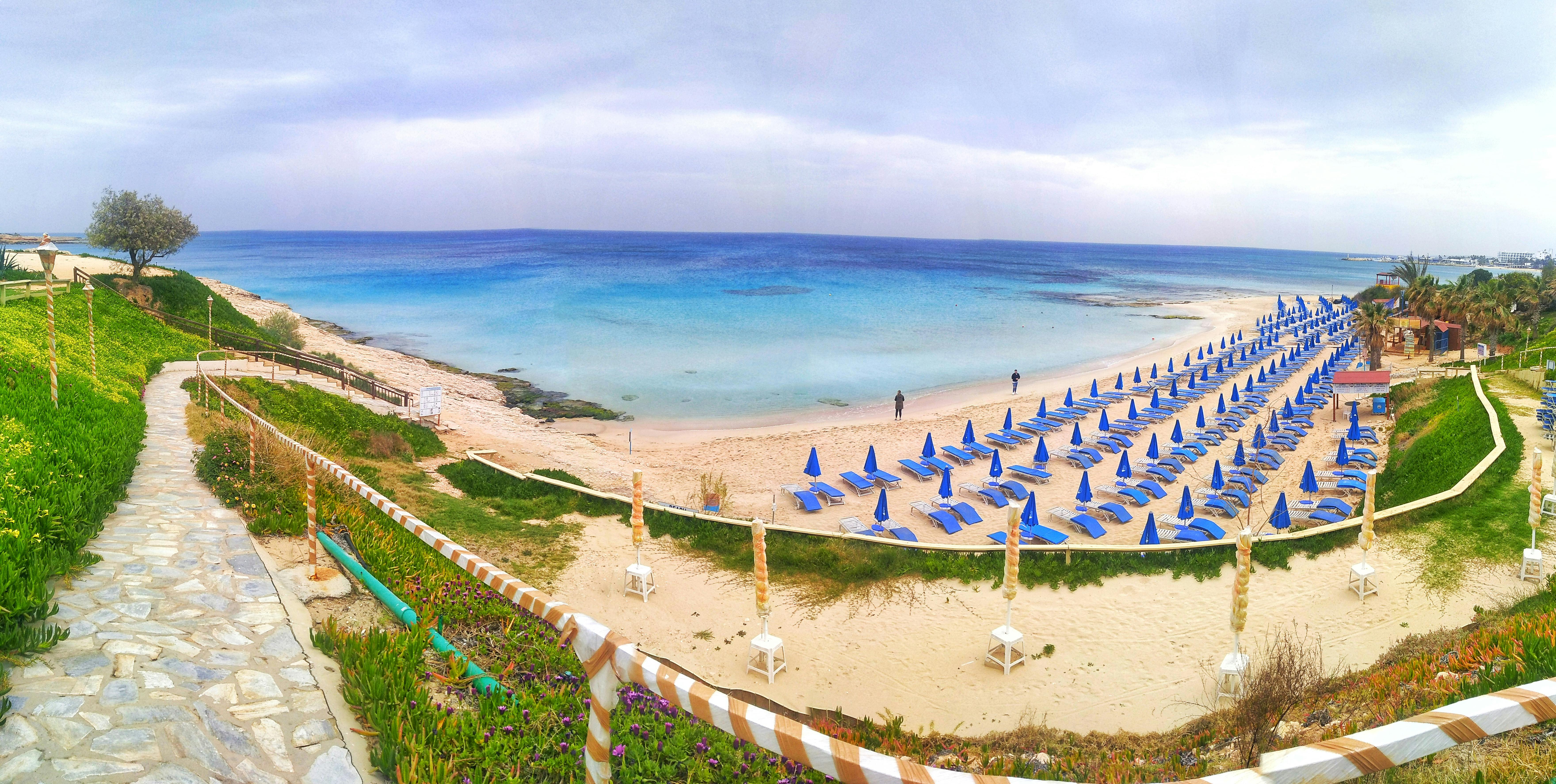 Picturesque panoramic view of colorful blue sea with horizon line near sandy shore with similar sun beds and growing grass with empty fenced pebble pathway under cloudy sky in summer