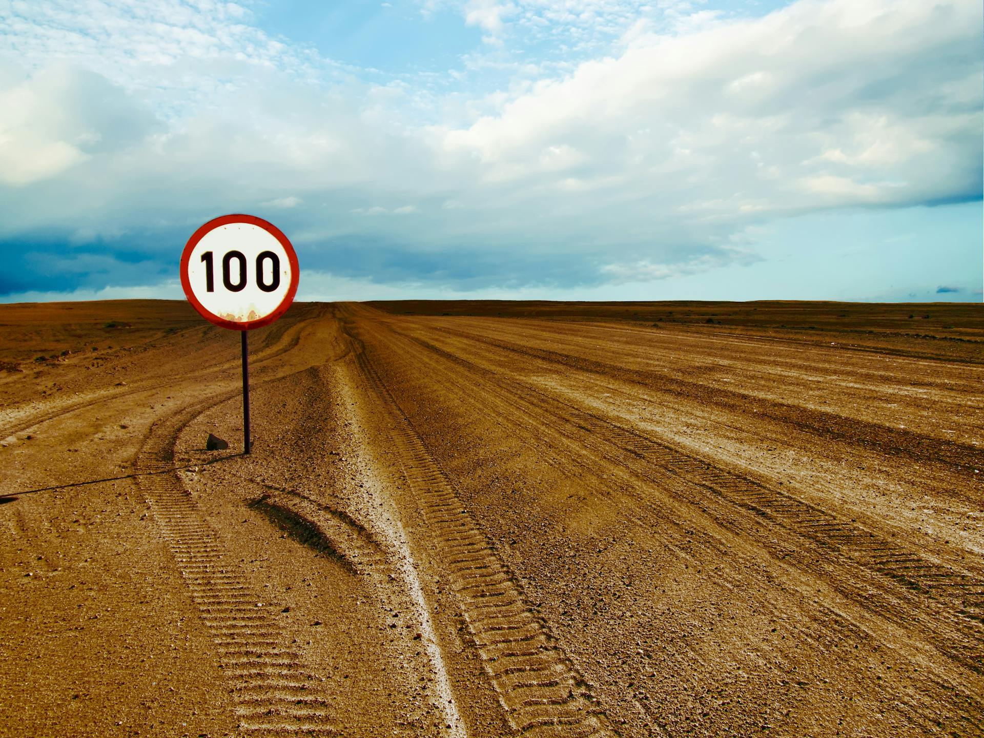 Expansive desert road with a visible 100 speed limit sign and cloudy sky.