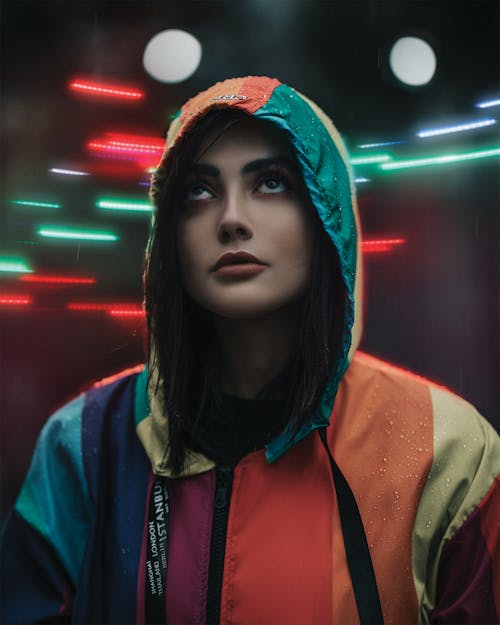 Young focused ethnic female in multicolored raincoat looking up while standing behind bright glowing lamps in evening in rainy weather