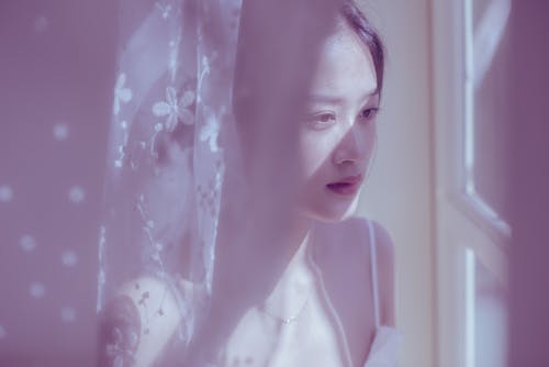 Tender young Asian female in top standing behind lace curtain and looking at window thoughtfully