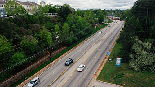 From above modern cars driving along asphalt road surrounded with green trees in suburb area