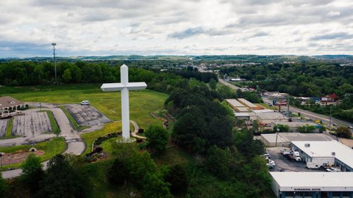 Free Drone view settlement with large white crucifix located on grassy lawn and surrounded with lush trees against commerce buildings Stock Photo