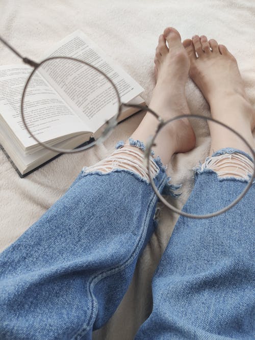 Crop faceless woman with eyeglasses reading book on bed