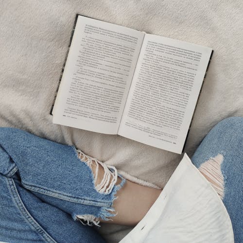 Top view crop anonymous female wearing casual outfit sitting on comfortable soft bed with legs crossed and reading good book in free time