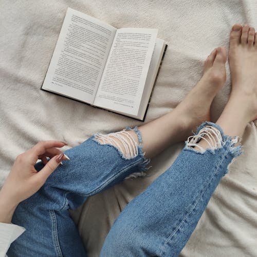 From above of crop anonymous barefooted female in trendy ripped jeans relaxing on bed and reading interesting book
