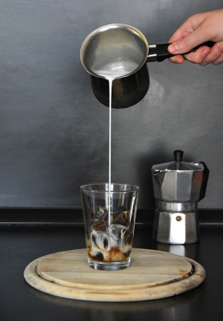 Milk being poured into iced coffee next to a moka pot on a kitchen counter.