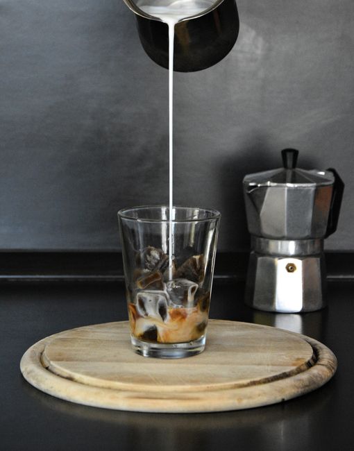 Milk pours into an iced coffee in a glass, beside a Moka pot, highlighting food preparation.