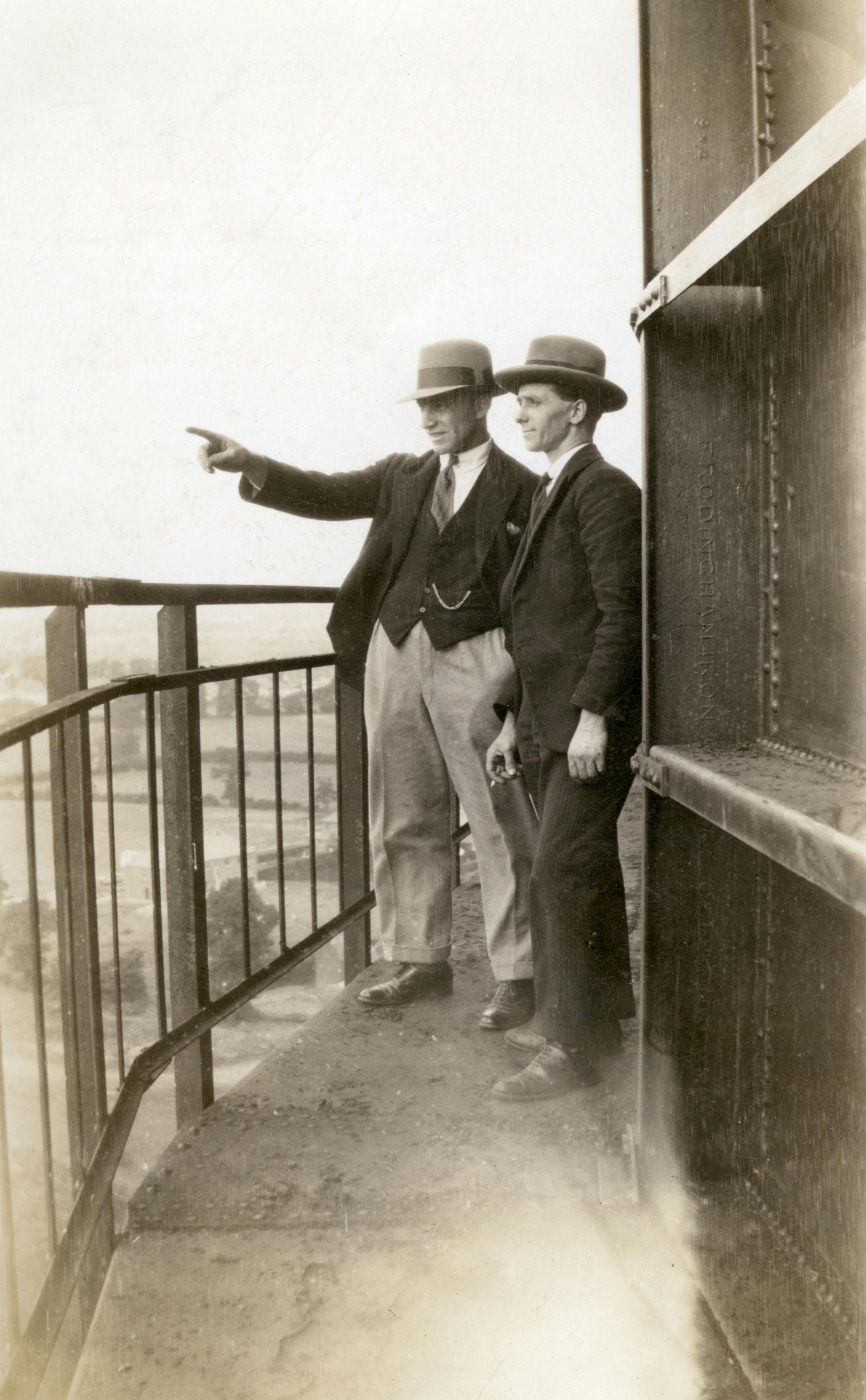Two men in vintage attire and hats stand on a balcony, pointing towards the distance.