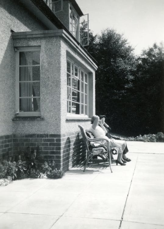 Grayscale Photo of People Sitting On A Bench Near Front Window Of A House