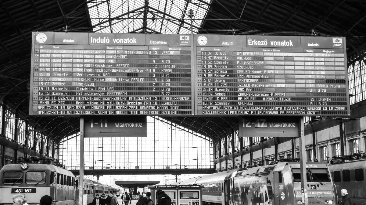 Black and white photo of a bustling train station with prominent departure board and commuters.