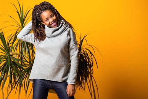 Cheerful young African American lady warm sweater touching long curly hair and smiling while standing near tropical plants against yellow background