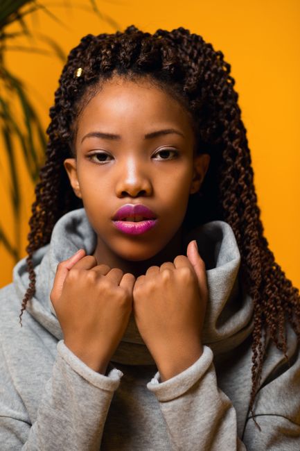 Young pensive ethnic female with long Afro hair touching trendy sweater collar and looking at camera against yellow background