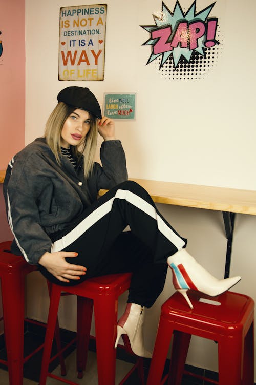 Full body of young self assured female in stylish outfit and cap sitting at counter on red stool near wall with creative stickers and looking at camera