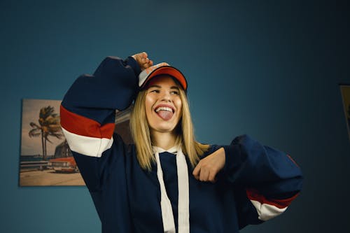 Playful young female in trendy outfit and cap showing tongue and smiling while having fun in modern studio