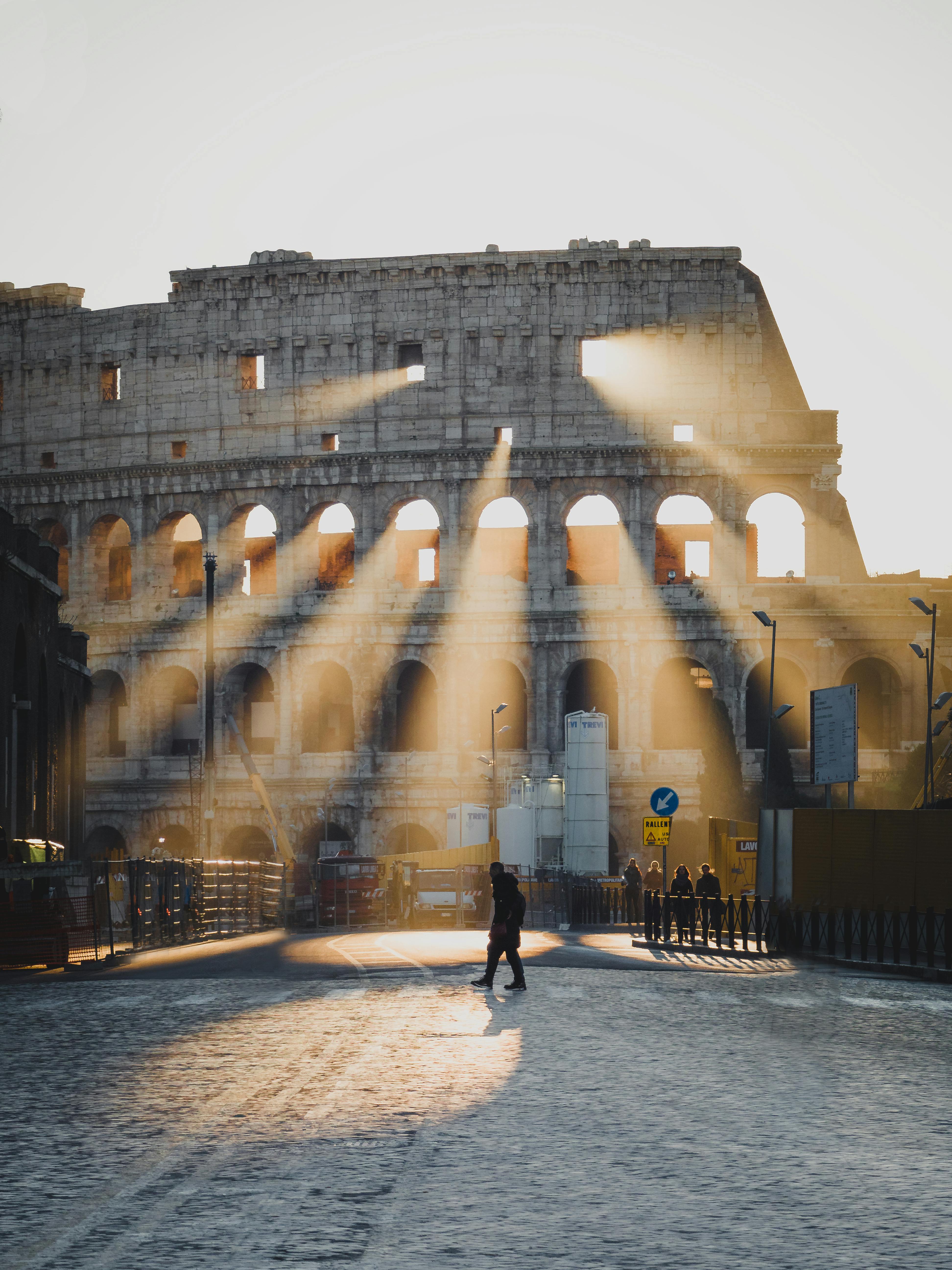 Free A person walking in front of the Colosseum in Rome, Italy, with sunrays filtering through the arches at sunrise. Stock Photo