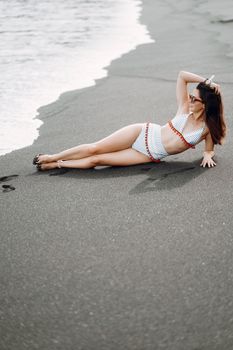 Full body barefoot female in swimwear and sunglasses lying on side on wet sandy beach near waving sea looking away