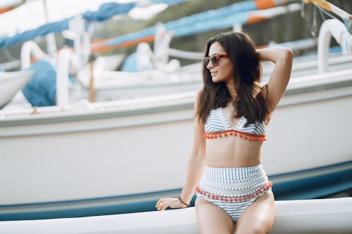 Optimistic female in charming swimwear and sunglasses sitting on yacht side on sea port near seashore while looking at sea