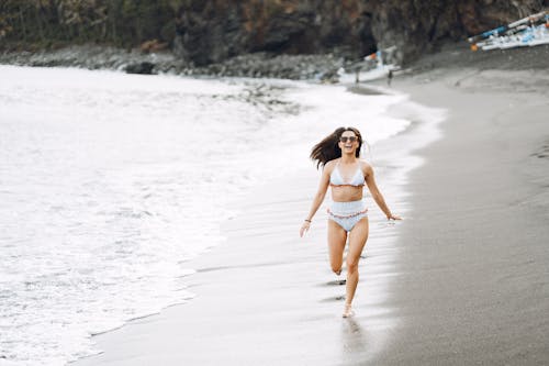 Free Full body happy slim female with loose hair in swimwear and sunglasses running along wet sandy coast near waving sea Stock Photo