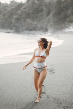 From above of cheerful young barefooted woman with long dark hair in swimsuit and sunglasses walking on sandy beach and looking at ocean while resting on seashore