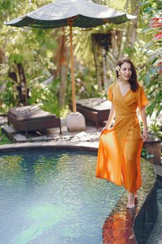 Full body barefoot positive female in orange summer maxi dress with flower in dark hair standing on poolside in tropical garden and looking away