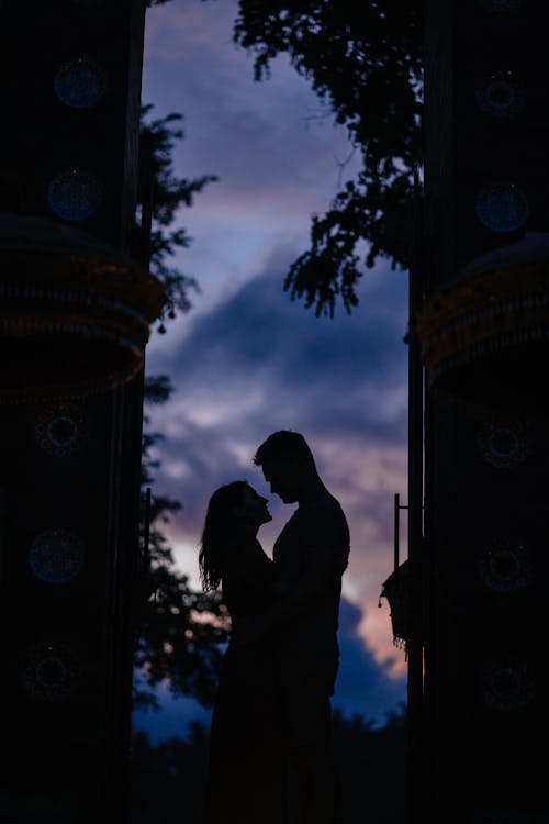 Side view unrecognizable silhouettes of bonding couple standing against dark blue sky in twilight and looking at each other near dark building walls