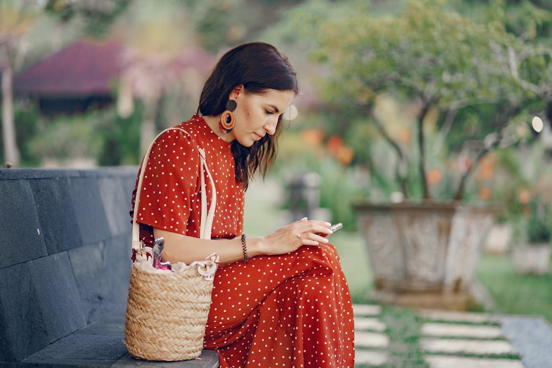 Side view of concentrated young female in trendy red summer dress with straw woven bag resting on bench in tropical park and browsing smartphone
