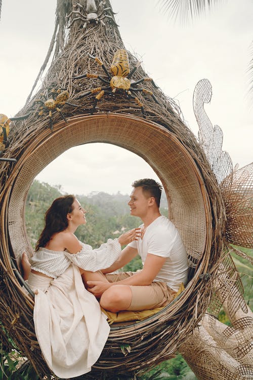 Side view full body barefoot content young couple in white casual clothes sitting in wicker hanging swing and looking at each other with love while spending honeymoon in tropical resort