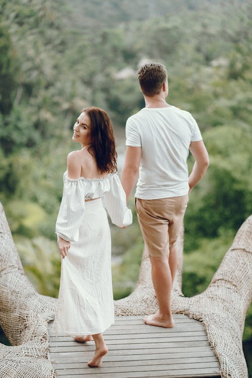 Free Back view full body cheerful couple in trendy white summer clothes standing on stone path and holding hands while enjoying scenic view of exotic tropics Stock Photo