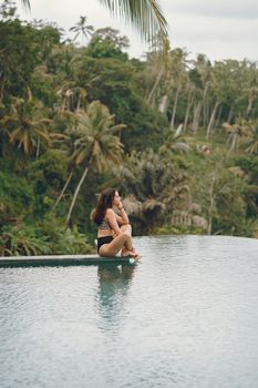 Side view full length anonymous thoughtful slim female in swimsuit sitting on poolside and enjoying amazing view of peaceful tropical landscape
