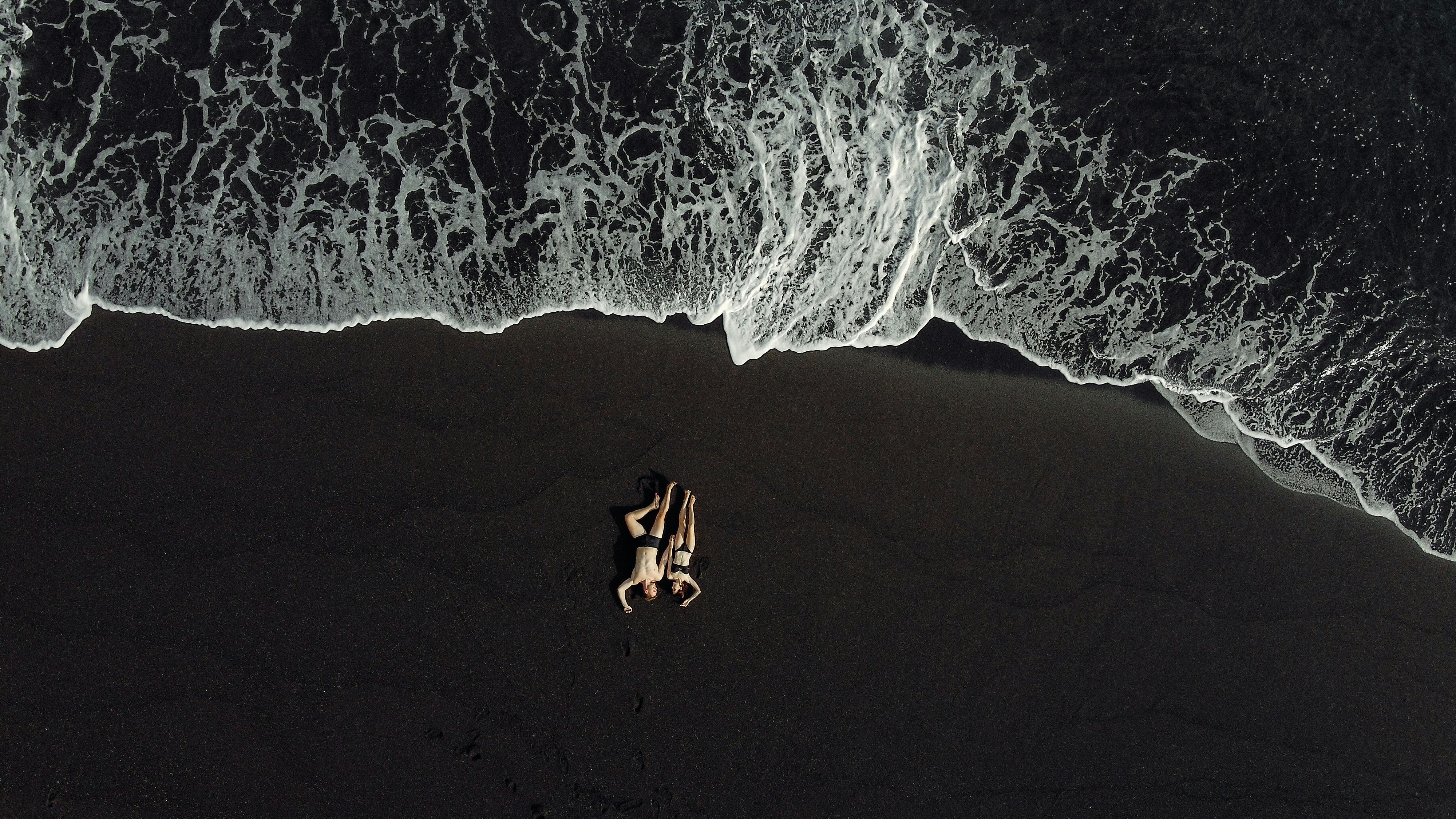 Drone view of relaxing hugging couple in dark swimwear chilling on black sand beach near splashing waving sea in daytime