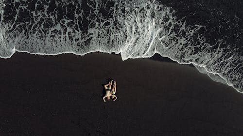 Drone view of relaxing hugging couple in dark swimwear chilling on black sand beach near splashing waving sea in daytime