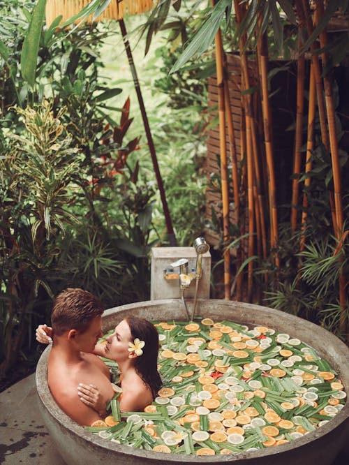 Side view from above of intimate couple hugging and looking at each other passionately while chilling in exotic stone bath with slices of orange and lemon in tropical spa