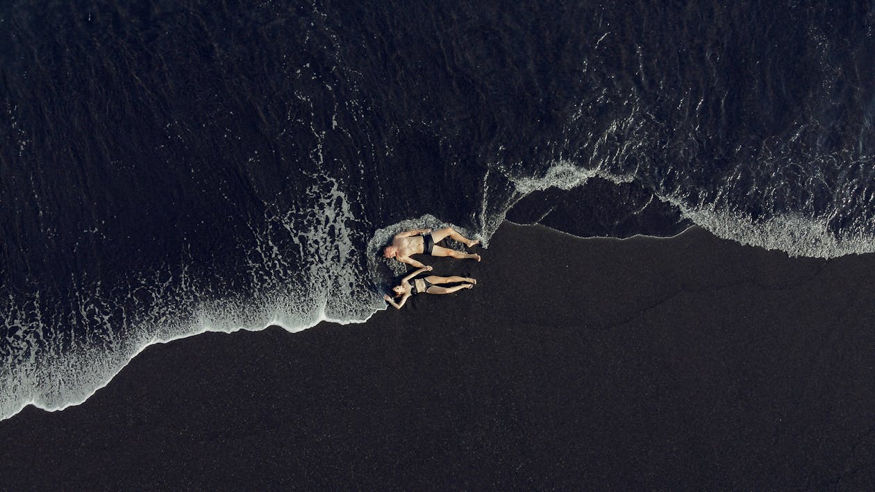 Drone view full body unrecognizable couple lying on black sand beach and holding hands near foamy sea waves in daylight