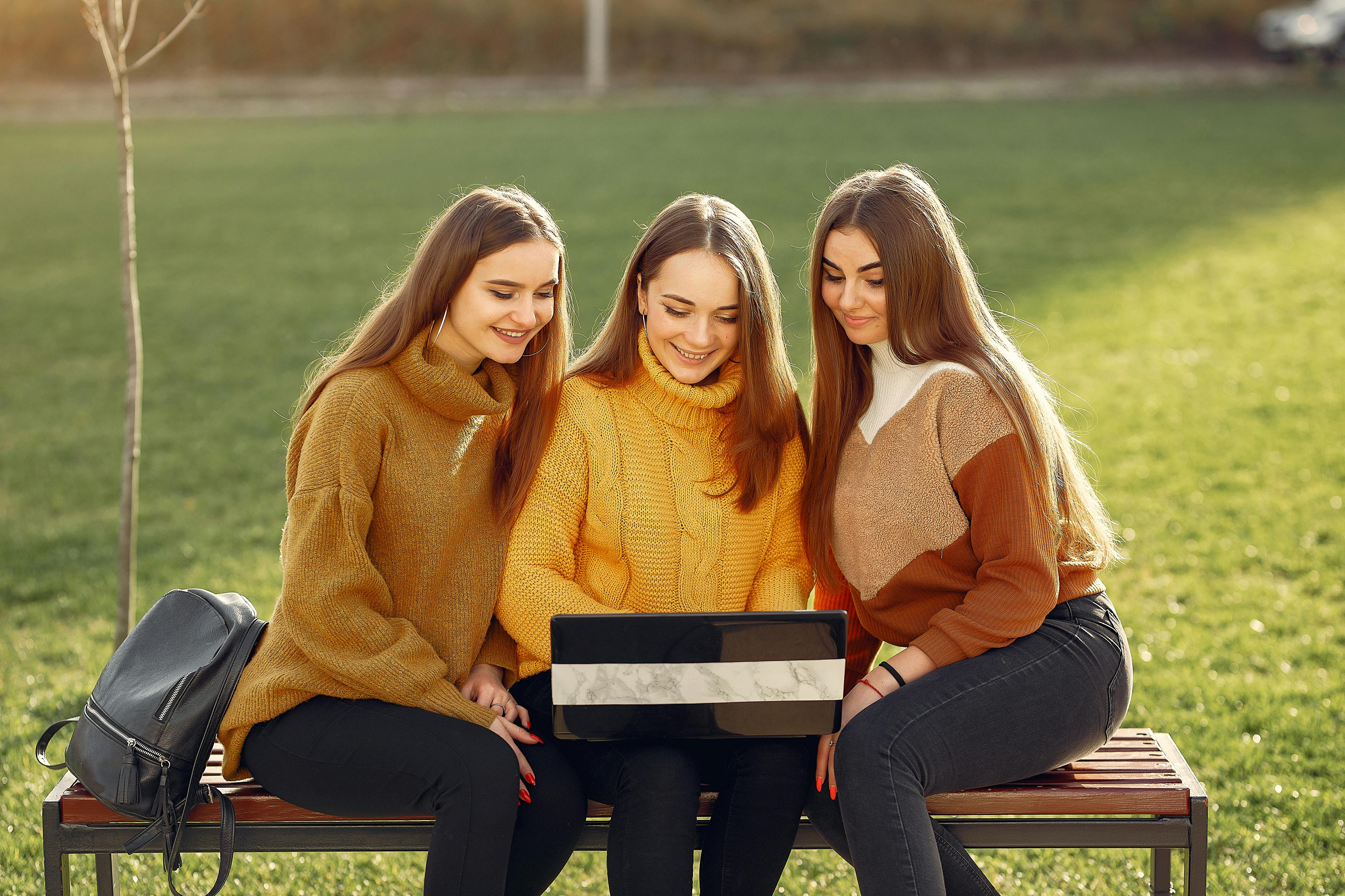 happy girlfriends using laptop while sitting on bench in park