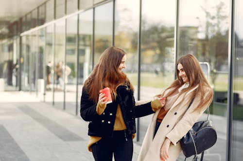 Young smiling best girlfriends in casual wear looking at each other and having fun while standing near glass building in city street