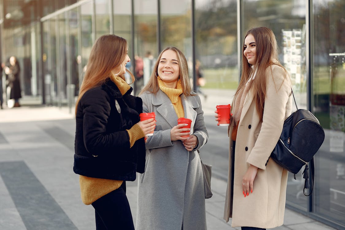 Beautiful female students standing near glass building while having friendly conversation outdoors